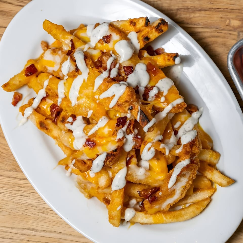 Loaded fries with melted cheese, bacon bits, and a drizzle of white sauce on a white plate.