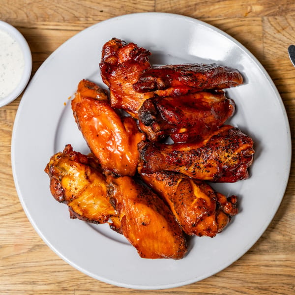A white plate filled with several glossy, saucy Brooklyn Wing Company chicken wings, with a side of ranch.